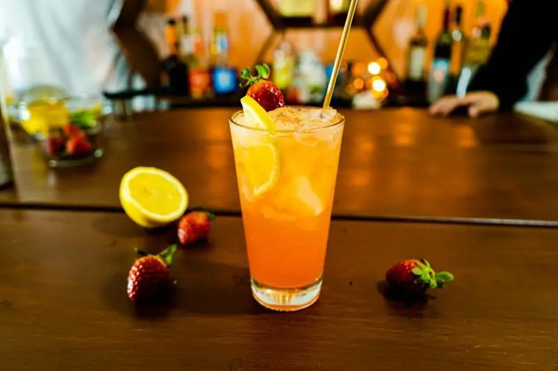 A cocktail on the table. Inside of cocktail some juice, liquor, lemon, strawberry and ice.