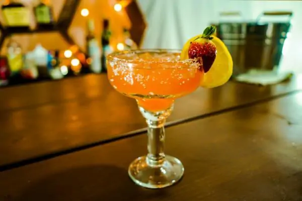 A pink colored cocktail in a big glass. It has strawberry and orange decoration, and rim salt around the glass